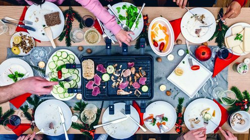 Vogelperspektive eines Esstischs mit Raclettegrill und Gem&uuml;se und Familie drumherum