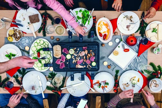 Vogelperspektive eines Esstischs mit Raclettegrill und Gemüse und Familie drumherum